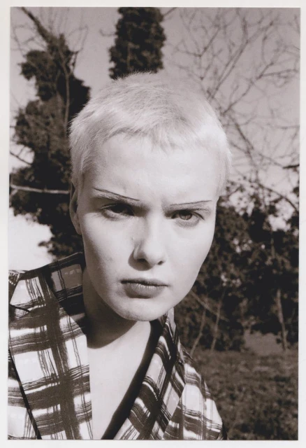 Portrait of an androgynous adult with short platinum hair, wearing a plaid jacket and striking a pose against an outdoor background of trees.