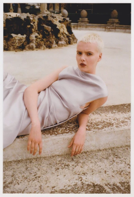 Elegant woman with short blonde hair wearing a grey dress posing outdoors near a stone fountain with a dark stone building in the background.