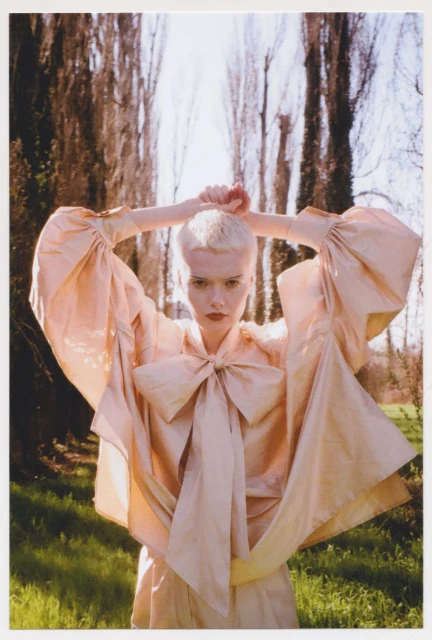 Fashion shot of a woman in a dress with a large bow, standing in a grassy field with tall trees under a bright sky, outdoor setting.