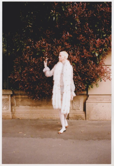 Elegant woman in a white fur stole poses on a city sidewalk against a backdrop of dark green and red-leaved bushes.