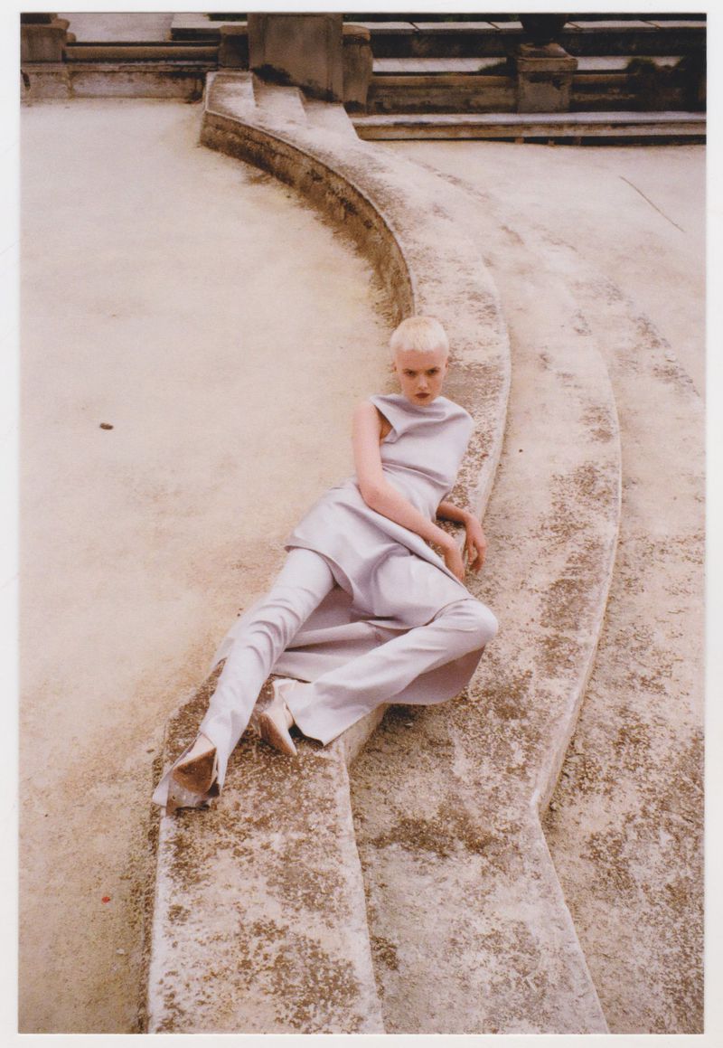Fashion photo of a model posing on concrete steps wearing light purple clothing and matching heeled shoes in a neutral-toned outdoor setting.