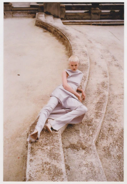 Fashion photo of a model posing on concrete steps wearing light purple clothing and matching heeled shoes in a neutral-toned outdoor setting.