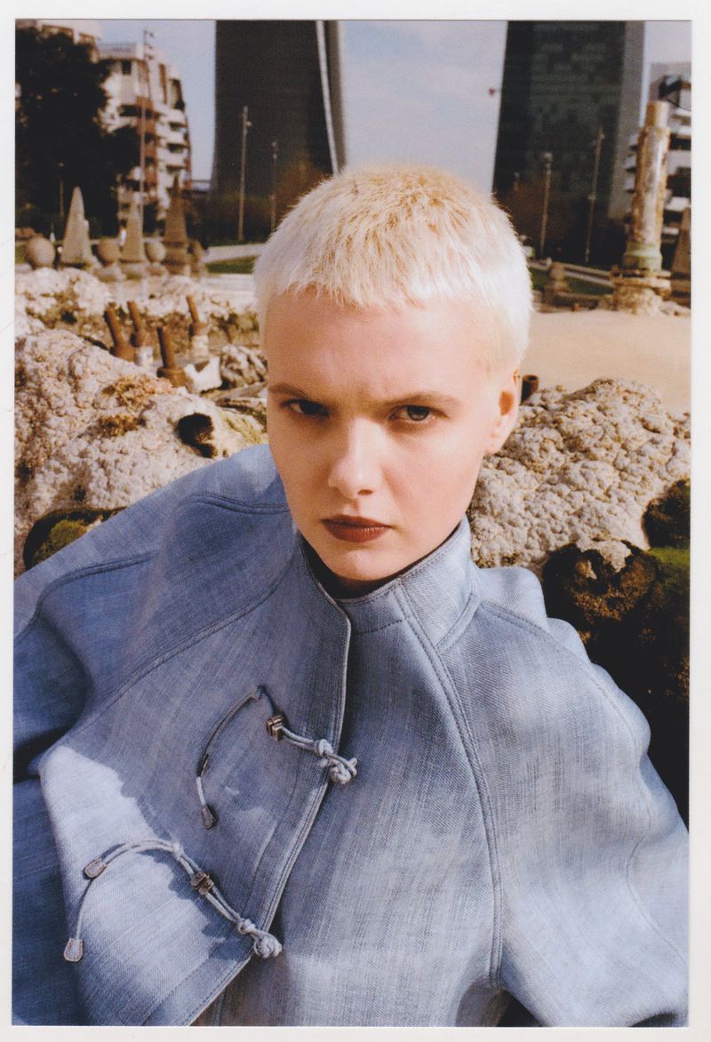 Woman with blonde hair in a blue jacket poses outdoors with modern architecture and stone structures in the background on a sunny day.