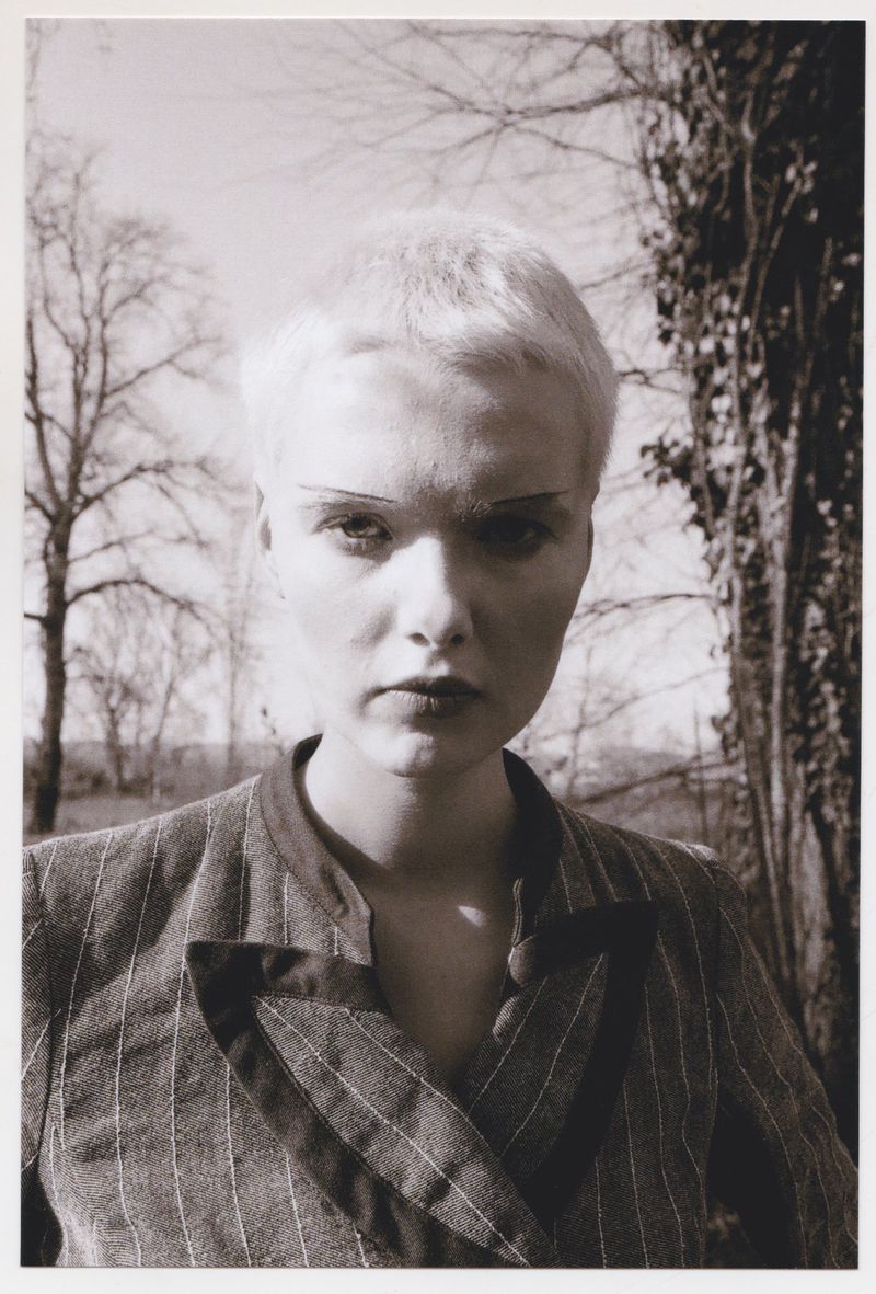 Close-up portrait of a model with short blonde hair wearing a pinstripe jacket outdoors against bare trees.