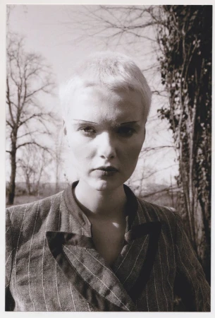 Close-up portrait of a model with short blonde hair wearing a pinstripe jacket outdoors against bare trees.