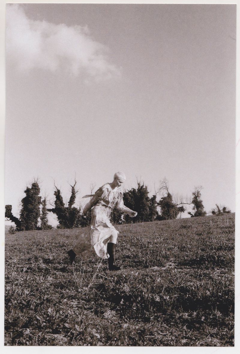 A young adult woman in a dress strides across a grassy hillside with trees in the distance beneath a cloudy sky, creating a natural scene.