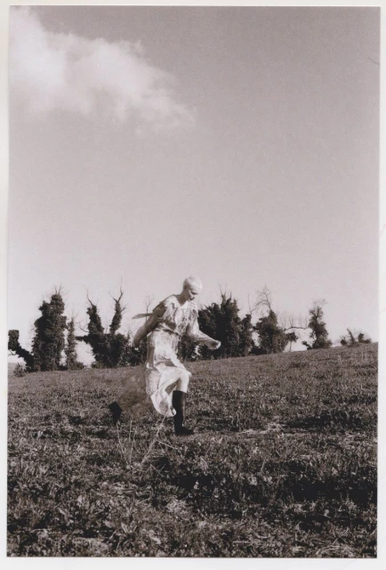 A young adult woman in a dress strides across a grassy hillside with trees in the distance beneath a cloudy sky, creating a natural scene.