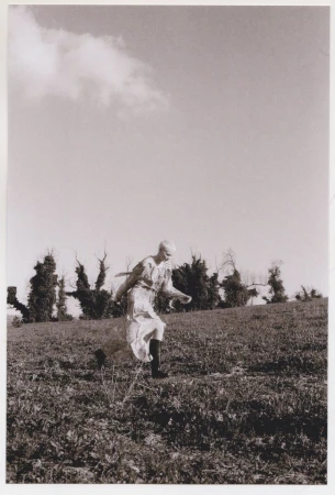 A young adult woman in a dress strides across a grassy hillside with trees in the distance beneath a cloudy sky, creating a natural scene.
