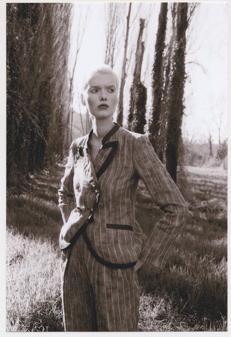 Monochrome portrait of a woman in a pinstripe suit, outdoors in a grassy field near tall trees, eyes gazing to the left side.