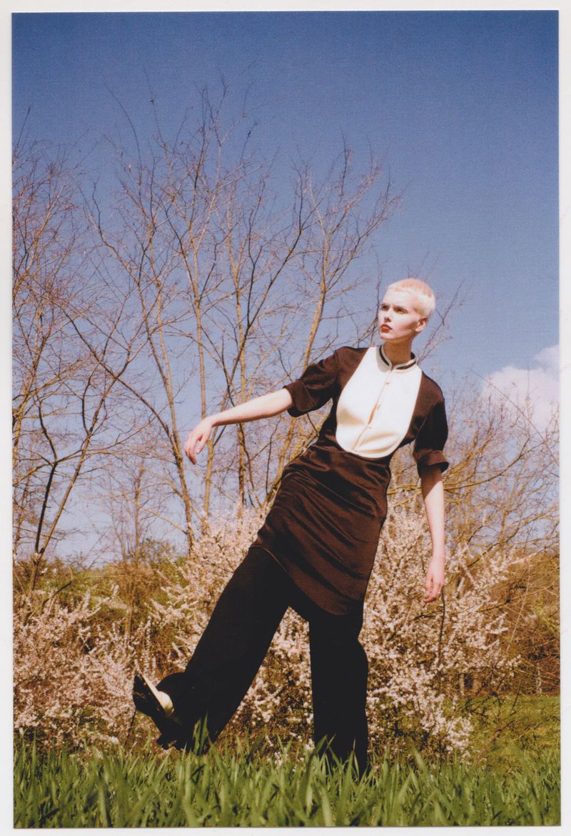 Short-haired model posing in a brown tunic and black trousers outdoors among blossoming trees against a clear blue sky.