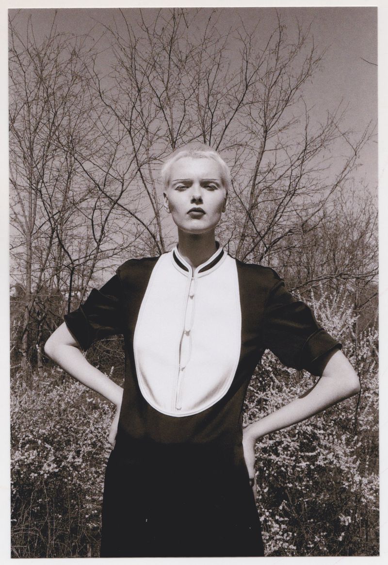 Monochrome portrait of an androgynous adult posing in nature wearing a dark dress with a white bib and bare trees in background.