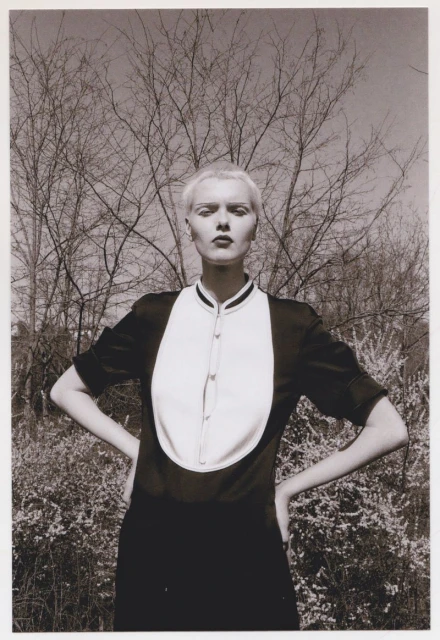 Monochrome portrait of an androgynous adult posing in nature wearing a dark dress with a white bib and bare trees in background.