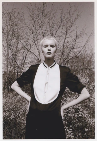 Monochrome portrait of an androgynous adult posing in nature wearing a dark dress with a white bib and bare trees in background.