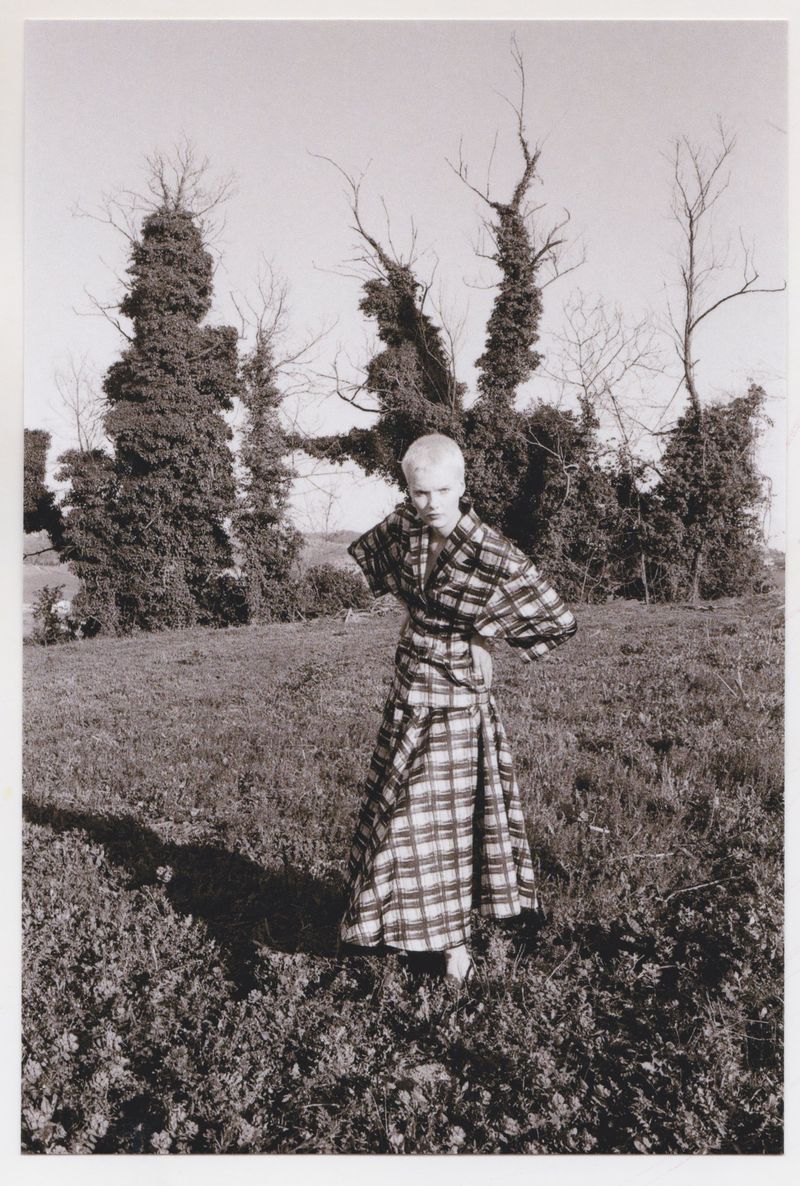 Adult female fashion model poses outdoors in a black-and-white plaid dress with hand-on-hip pose in a grassy field near trees.