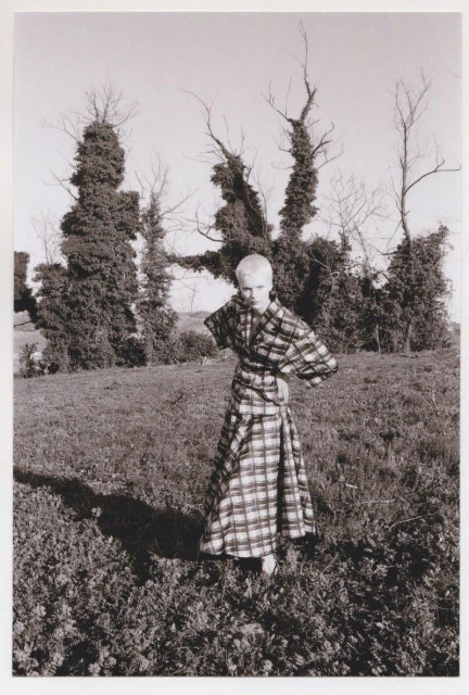 Adult female fashion model poses outdoors in a black-and-white plaid dress with hand-on-hip pose in a grassy field near trees.