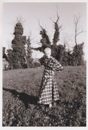 Adult female fashion model poses outdoors in a black-and-white plaid dress with hand-on-hip pose in a grassy field near trees.