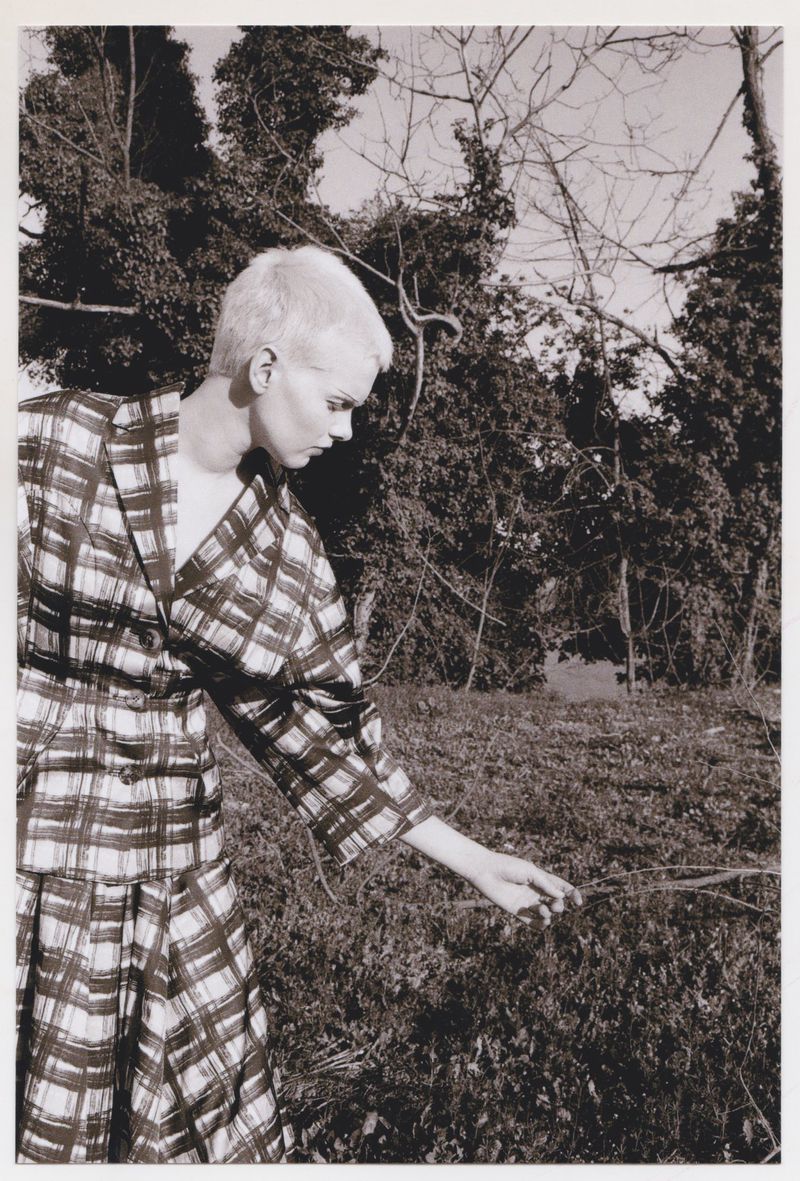 Monochrome fashion shot of a woman in a plaid outfit in a grassy field with a forest backdrop, creating a vintage aesthetic.