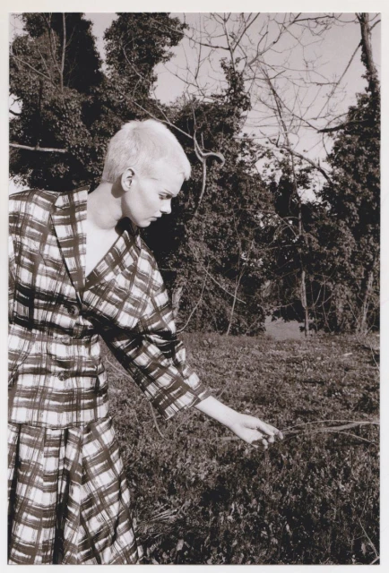 Monochrome fashion shot of a woman in a plaid outfit in a grassy field with a forest backdrop, creating a vintage aesthetic.