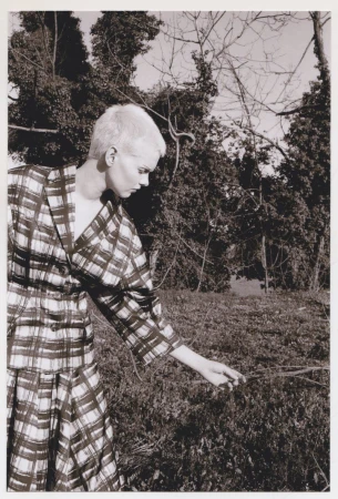 Monochrome fashion shot of a woman in a plaid outfit in a grassy field with a forest backdrop, creating a vintage aesthetic.