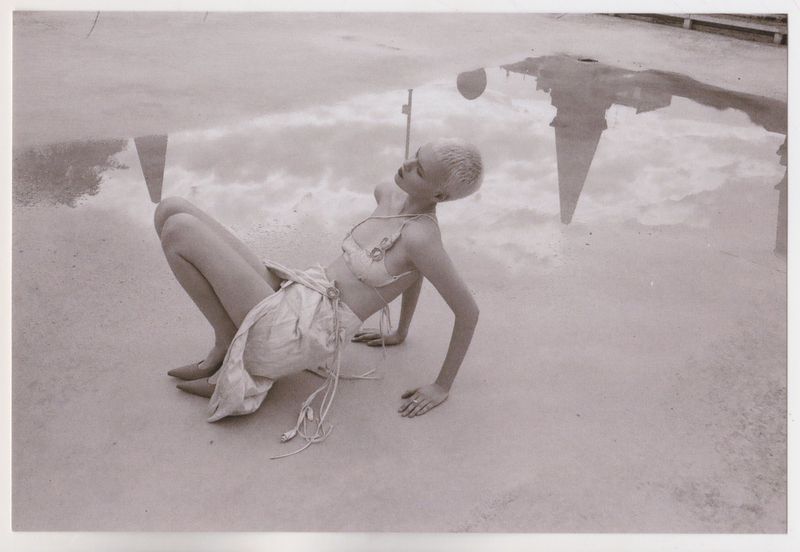 Model with short platinum blonde hair posing near a large puddle reflecting building spires and sky, captured in a sepia-toned editorial.