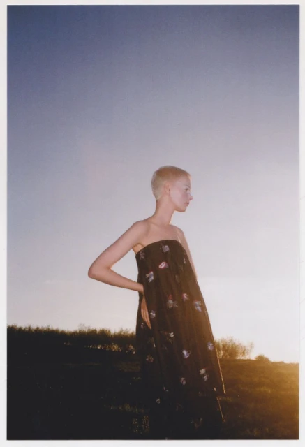 Young woman with short blond hair and a dark floral dress stands outdoors against a background of setting sun and blue sky.
