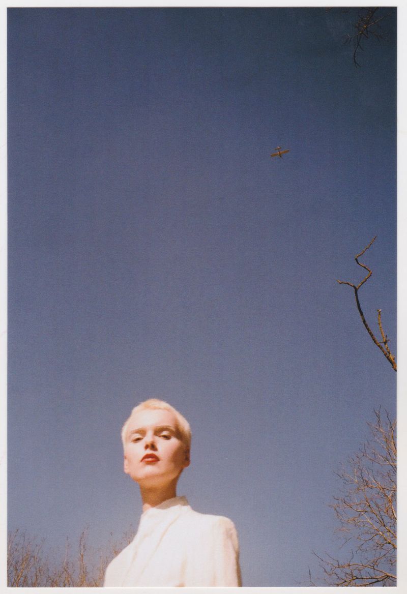 Adult female with short blonde hair and red lipstick looking up towards a blue sky with a small plane and tree branches.