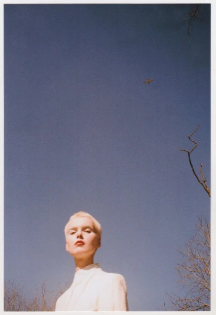 Adult female with short blonde hair and red lipstick looking up towards a blue sky with a small plane and tree branches.