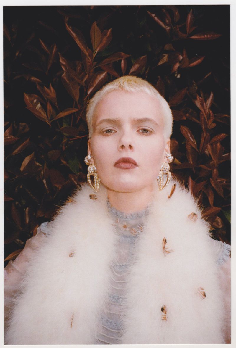 Model with short blonde hair wearing ornate earrings and a feathered white collar against dark foliage