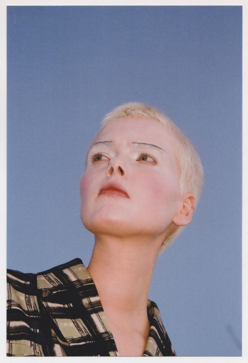 Low-angle shot of a fair-skinned adult woman with short, blonde hair and white eyebrows, wearing a green and black checkered shirt against a blue sky.