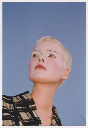 Low-angle shot of a fair-skinned adult woman with short, blonde hair and white eyebrows, wearing a green and black checkered shirt against a blue sky.