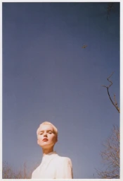 Model with short blonde hair and red lipstick wearing white top against blue sky with distant airplane and bare tree branches