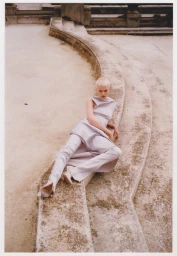 Model with short blonde hair in a light grey outfit reclining on curved stone steps outdoors.