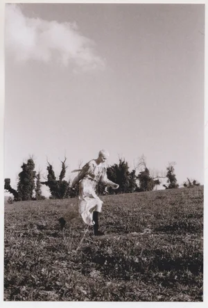 Model in patterned dress and dark boots walking across a sloping grassy field under an open sky.
