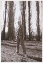 Model with short blonde hair wearing a patterned suit standing outdoors in a grassy field surrounded by tall bare trees.