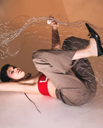 A woman lies on a white surface with her legs up, posing in a red top, grey pants, and black high heels while holding up a wire mesh.