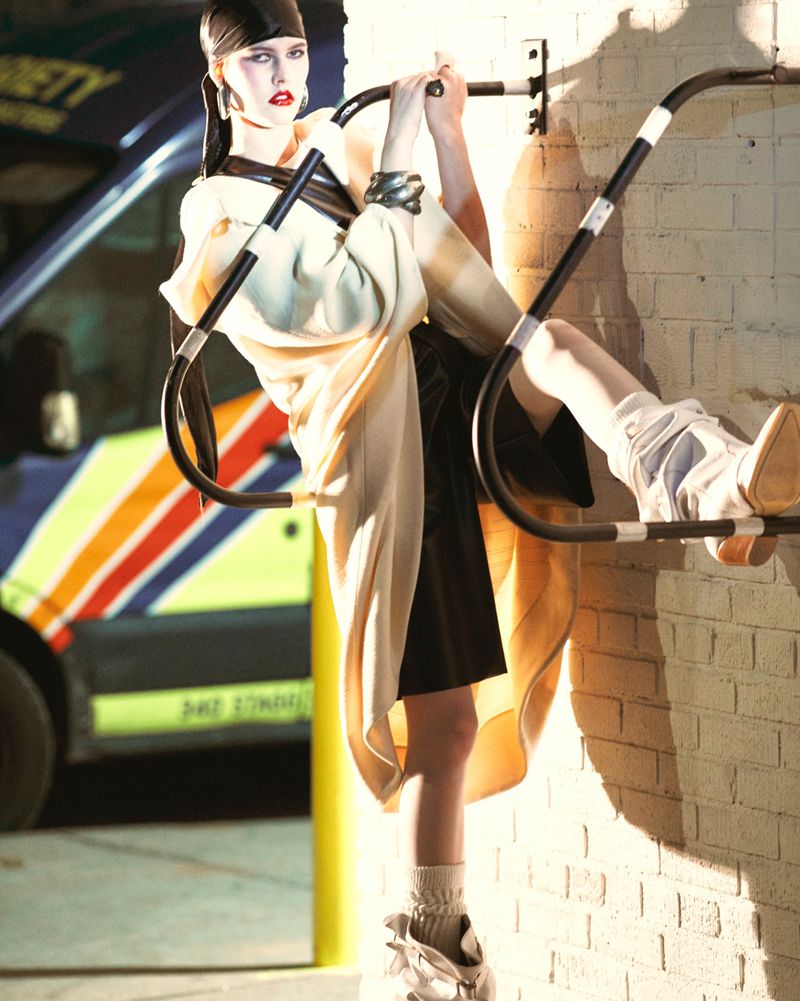 Fashionable woman in avant-garde outfit poses on metal bars in front of a commercial van and brick wall with directional lighting.