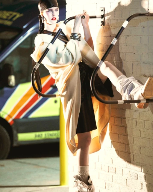 Fashionable woman in avant-garde outfit poses on metal bars in front of a commercial van and brick wall with directional lighting.