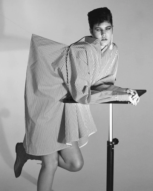 Monochrome portrait of a young adult woman in striped oversized shirt-dress, leaning on an adjustable table in a light grey studio.