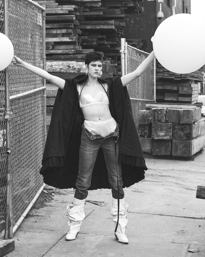 Monochrome fashion shot of a woman with short hair wearing jeans, lingerie, and a cape, holding white balloons outdoors.