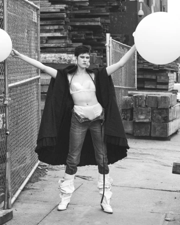 Monochrome fashion shot of a woman with short hair wearing jeans, lingerie, and a cape, holding white balloons outdoors.