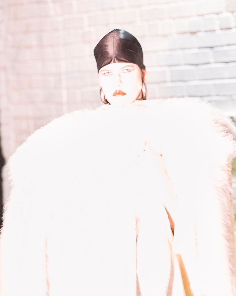 A person in a white faux fur coat and black head covering stands in front of a grey brick wall, looking directly at the camera.