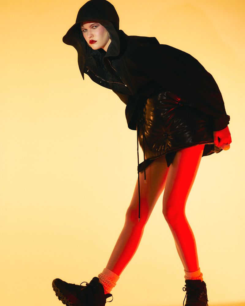 Model in black hooded jacket and skirt with bright red tights against a warm yellow gradient studio backdrop.