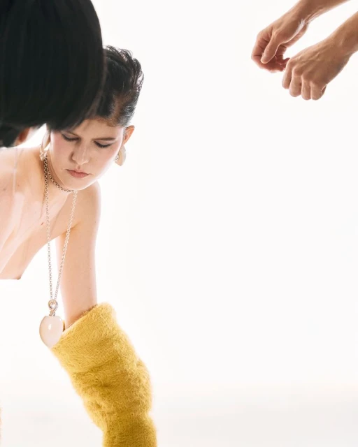 Indoor fashion shoot features a woman with dark hair and a fair complexion, wearing a white top, necklace, and yellow arm covering.