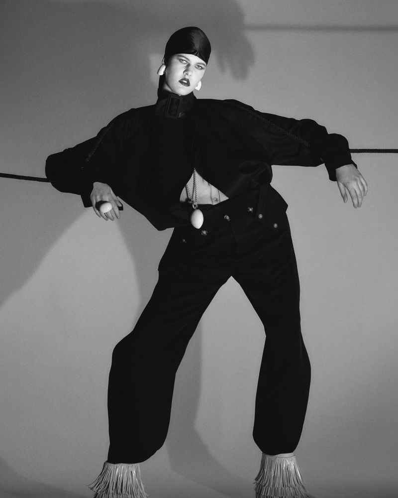 Black and white fashion shot of a person with dark hair in a black outfit, leaning on a black line in a light grey studio.