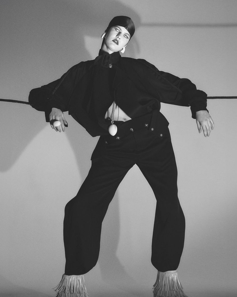Monochrome fashion shot of a person in a dark outfit with avant-garde styling and striking accessories, posing in a studio setting.