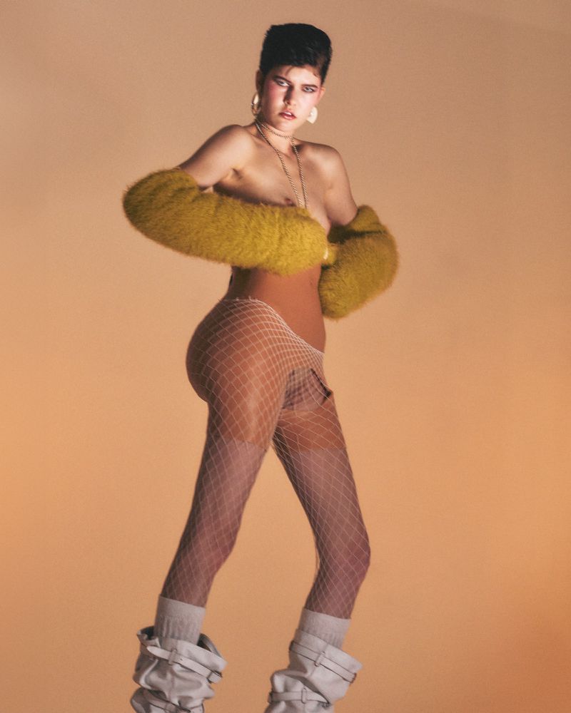A fashion portrait of a woman in leg warmers and fur sleeves against a gradient orange background in a studio setting.