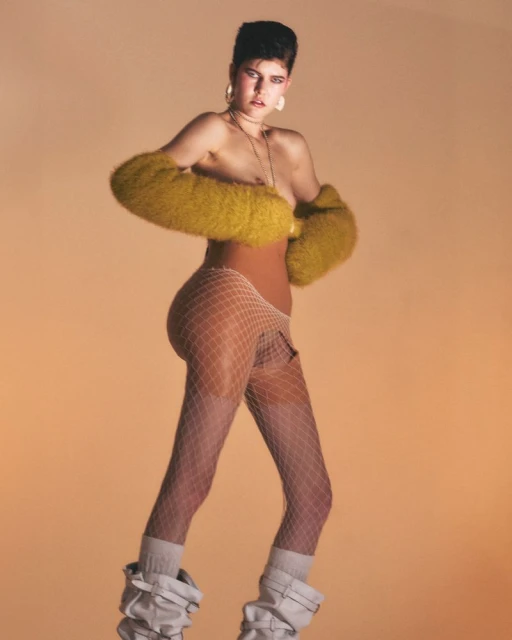 A fashion portrait of a woman in leg warmers and fur sleeves against a gradient orange background in a studio setting. 