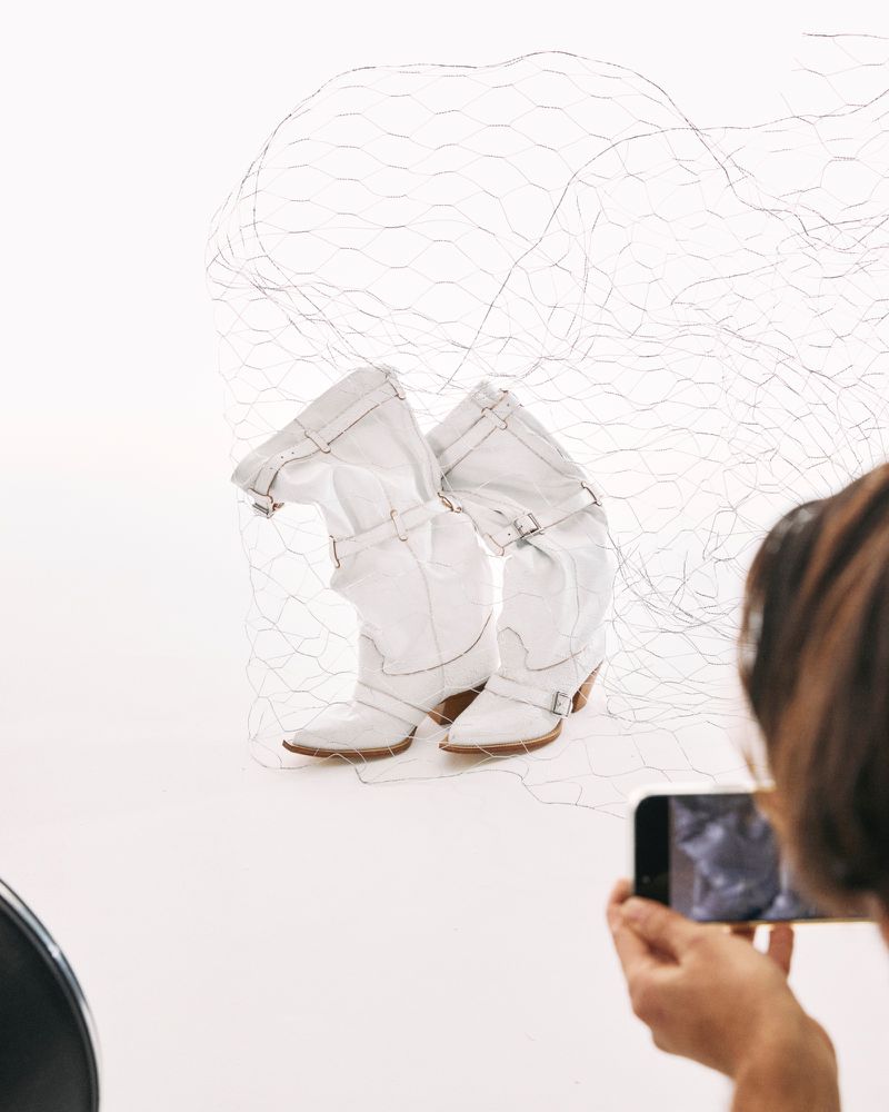 White heeled cowboy boots are photographed in a photo studio by a person using their smartphone, with a wire mesh in the background.