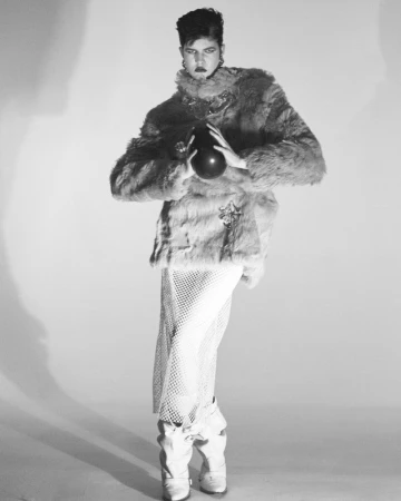 Model Rachel Goff in a fuzzy jacket and mesh skirt holding a dark object against a seamless studio background.