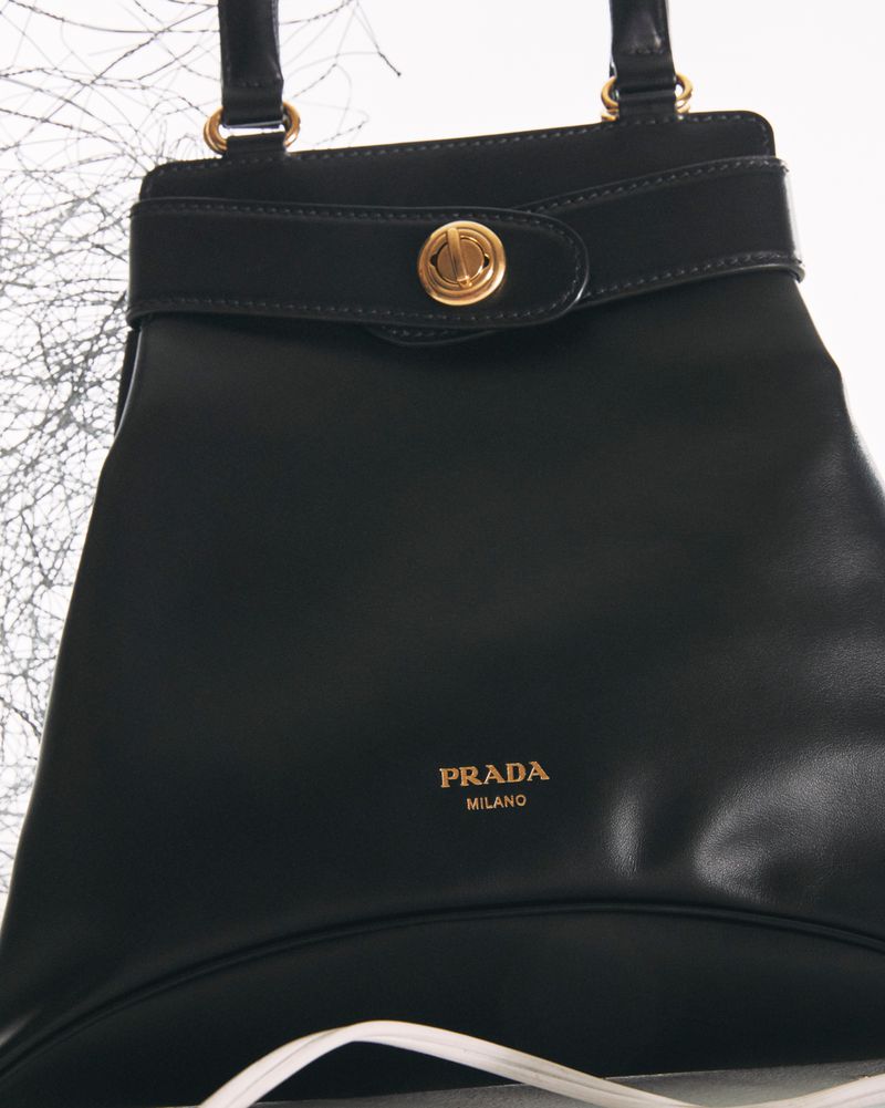 Close-up of a black Prada handbag featuring golden lettering and hardware, set against a blurred background.